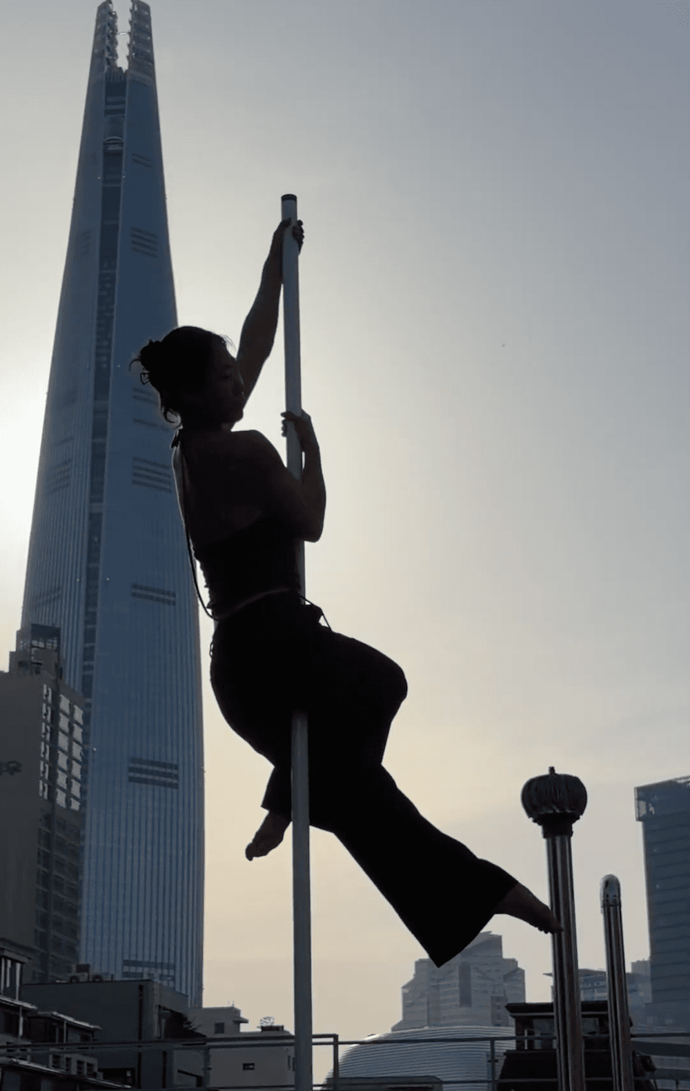 Haeri doing a pole dance trick on a rooftop with Jamsil tower and the Seoul City skyline in the background.